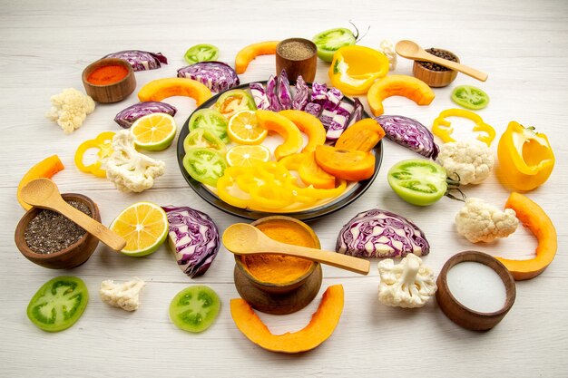 Bottom view chopped vegetables and fruits pumpkin persimmon red cabbage lemon green tomatoes cauliflower yellow bell peppers on round platter spices in small bowls on white table