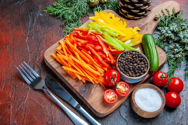 Bottom view colorful cut peppers black pepper tomatoes cucumber on cutting board pine branches salt fork and knife on dark red table