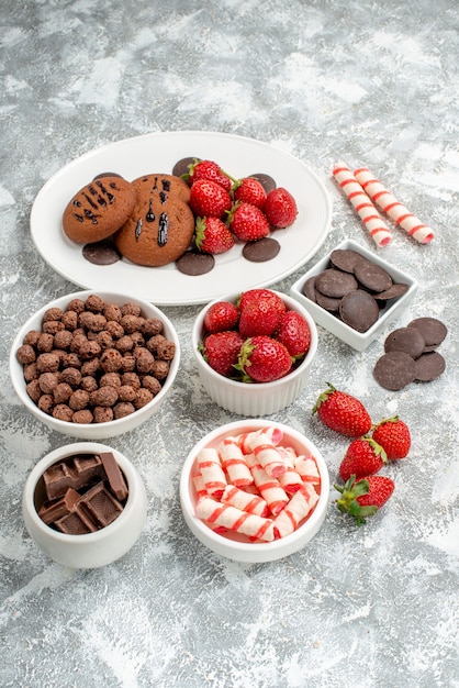Вид снизу печенье клубника и круглые шоколадные конфеты на белой овальной тарелке, тарелки с конфетами клубника, шоколадные хлопья на серо-белом столе