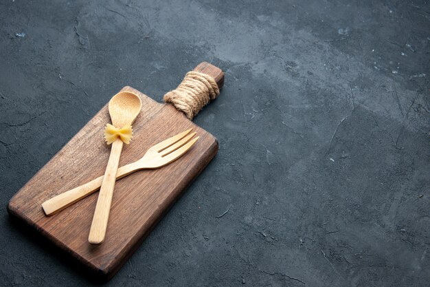 Free photo bottom view crossed wooden spoon and fork on wooden serving board on dark surface with free space