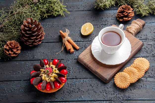 Bottom view a cup of tea on chopping board slice of lemon pinecones cookies cinnamon and berry cake on dark wooden background