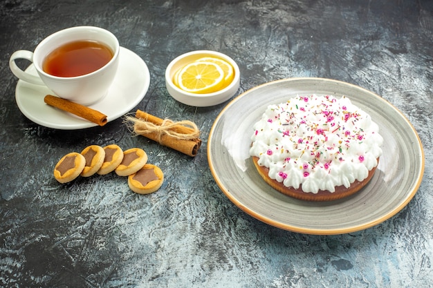 Vista dal basso, tazza di tè, fette di limone in una piccola torta con piattino su un piatto rotondo, biscotti, bastoncini di cannella sul tavolo scuro