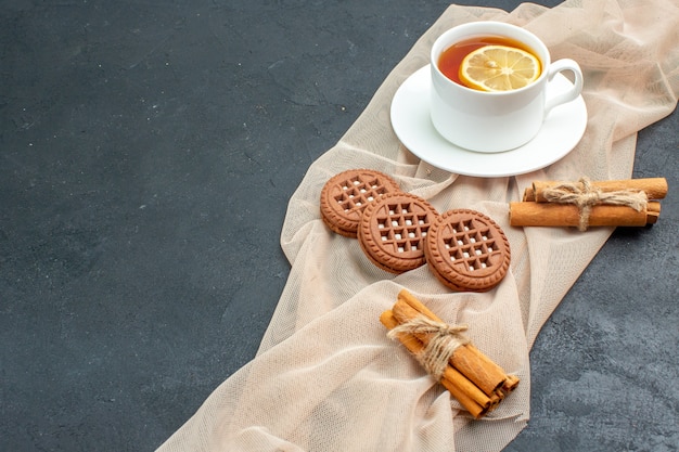 Вид снизу чашка чая с лимоном, палочками корицы, печеньем на бежевой шали на темной поверхности, свободное пространство