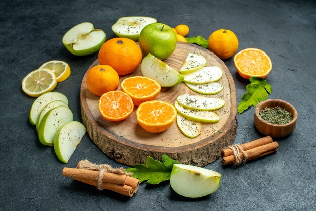 Bottom view cut apples and mandarines on round wood board cinnamons tied with rope dried mint powder in small bowl on dark table