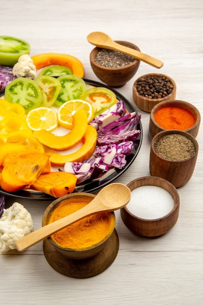 Bottom view cut vegetables and fruits pumpkin bell peppers persimmon red cabbage on black plate spice in small bowls wooden spoon on wooden table