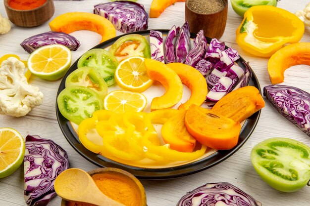 Bottom view cut vegetables and fruits pumpkin persimmon red cabbage lemon green tomatoes cauliflower bell peppers on round platter various spices in small bowls on table