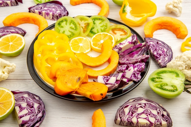 Bottom view cut vegetables and fruits pumpkin persimmon yellow bell peppers red cabbage lemon green tomatoes on black round platter on white table