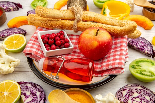 Вид снизу ягоды шиповника в миске яблочный хлеб красная бутылка на салфетке на круглой тарелке нарезанные овощи на белом столе