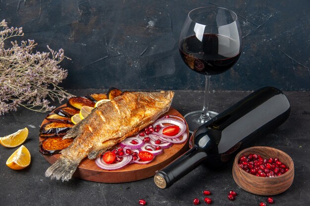 Вид снизу, жареная рыба, жареные баклажаны, нарезанный лук на деревянной доске, бутылка вина, лежа и бокал на темном фоне
