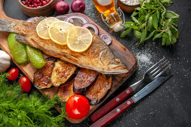 Bottom view fish fry fried eggplants onion peppers on wood board spices in small bowls fork and knife tomatoes oil bottle mint dill on dark background