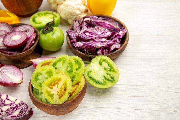 Bottom view fresh vegetables cut onion cut green tomatoes cut red cabbage in bowls bell pepper cauliflower on white wooden table