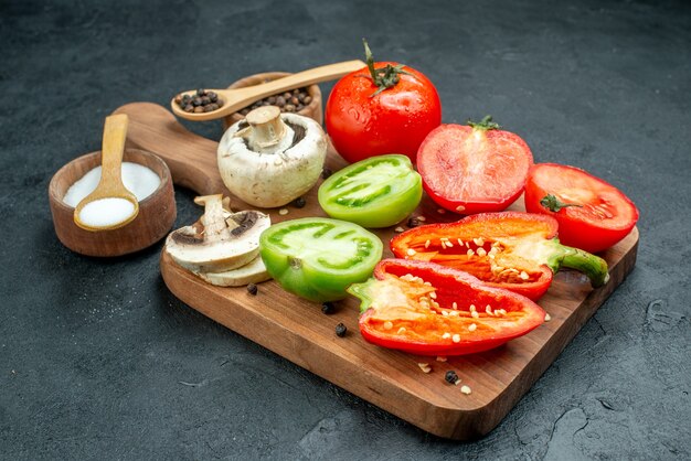 Bottom view fresh vegetables mushrooms cut red and green tomatoes bell peppers on chopping board bowls with black pepper and salt wooden spoons on dark table