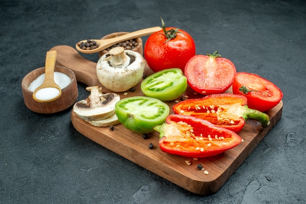 Вид снизу свежие овощи грибы нарезанные красные и зеленые помидоры болгарский перец на разделочной доске миски с черным перцем и солью деревянные ложки на темном столе