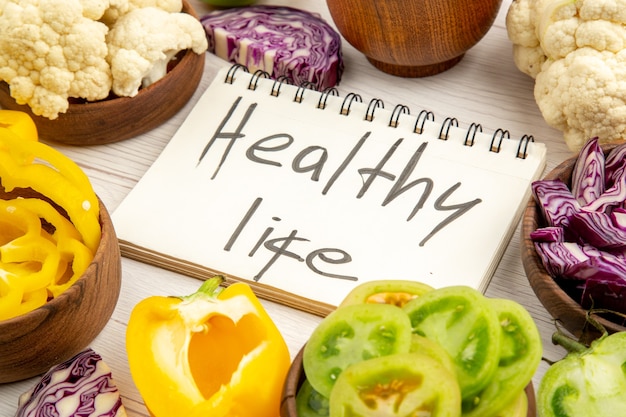 Bottom view healthy life written on notebook red cabbage cauliflower yellow bell pepper green tomato in bowls on white wooden surface