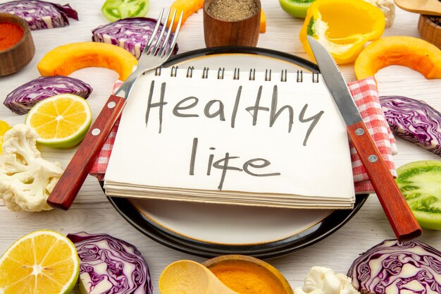 Bottom view healthy life written on notepad fork and knife on round plate cut vegetables on white table