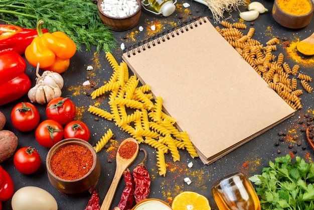 Foto gratuita vista dal basso delle spezie di verdure fresche del blocco note della pasta a spirale cruda sulla tavola nera