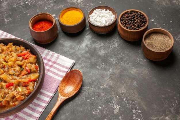 Bottom view roasted eggplant salad in bowl wooden spoon different spices in bowls on dark table