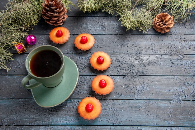 Bottom view rounded cherry cupcakes fir-tree branches christmas toys cones and a cup of tea on dark wooden background