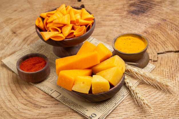 Bottom view slices of cheese in bowl turmeric red pepper powder chips wheat spike on newspaper on wooden table