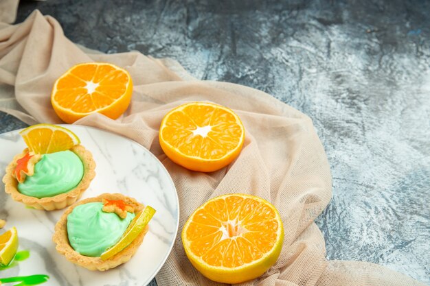 Bottom view small tarts with green pastry cream and lemon slice on plate beige shawl cut oranges on dark surface with free space