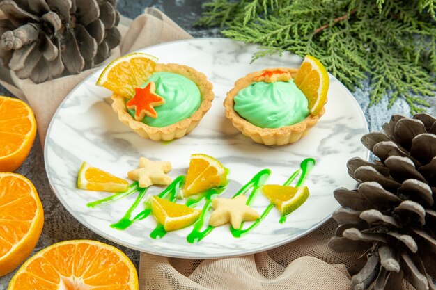 Bottom view small tarts with green pastry cream and lemon slice on plate on beige shawl cut oranges on dark surface