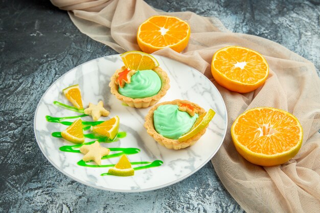 Bottom view small tarts with green pastry cream and lemon slice on plate on beige shawl cut oranges on dark surface