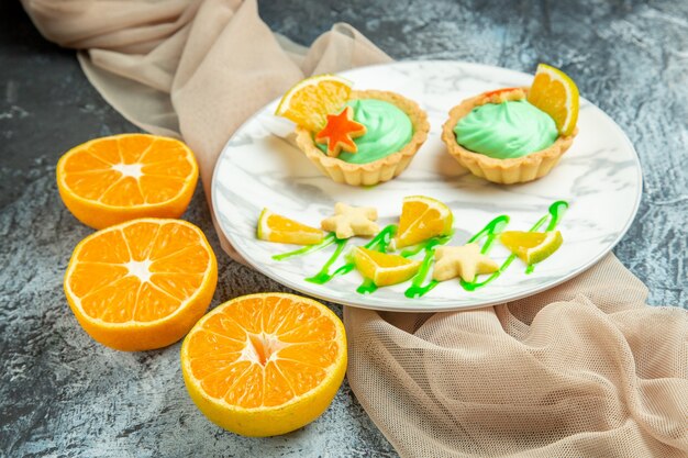 Bottom view small tarts with green pastry cream and lemon slice on plate on beige shawl cut oranges on dark surface