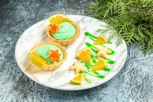 Bottom view small tarts with green pastry cream and lemon slice on plate pine branch on dark surface