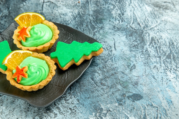 Bottom view small tarts with green pastry cream xmas tree biscuits on black plate on grey surface with copy space