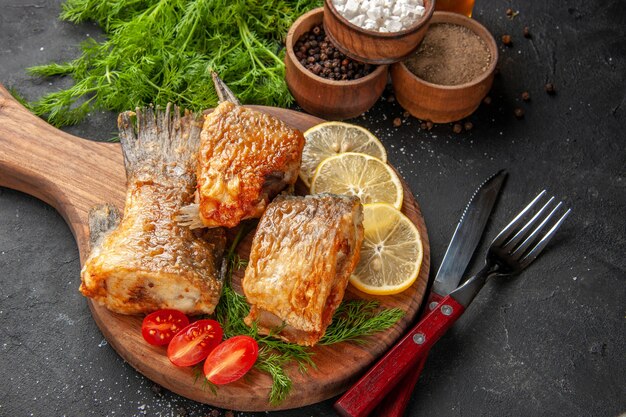 底面図おいしい魚のフライレモンスライスはまな板でチェリートマトをカットボウルナイフとフォークで黒の背景にさまざまなスパイス