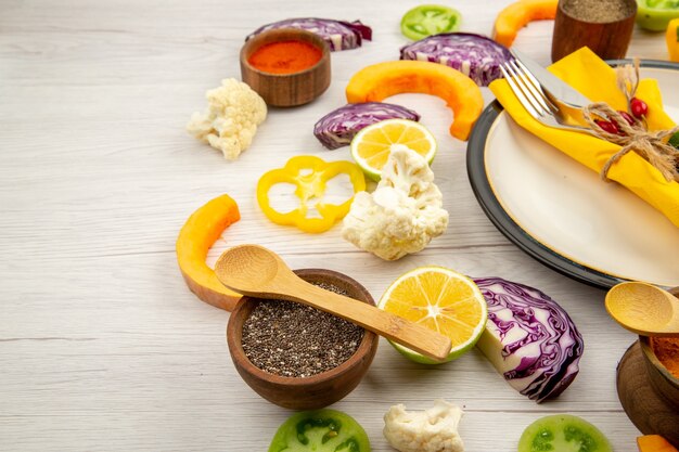 Bottom view tied knife and fork with yellow napkin on white platter cut vegetables red cabbage pumpkin cauliflower yellow bell pepper spices in small bowls on white wooden table