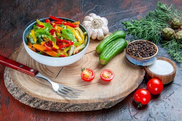 Bottom view vegetable salad in bowl fork garlic black pepper cucumbers on rustic board fir branch on dark red table