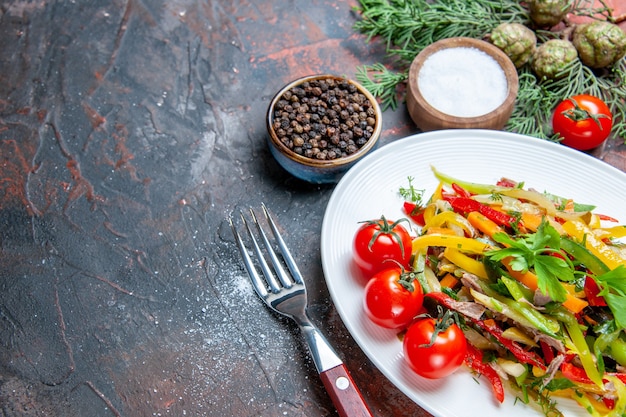 Вид снизу овощной салат на овальной тарелке помидоры черри вилка черный перец и соль на темно-красном столе свободное пространство