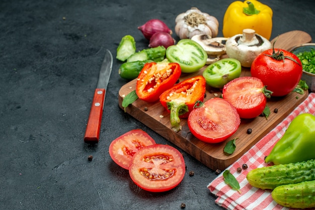 Bottom view vegetables green and red tomatoes bell peppers on cutting board greens in bowl knife cucumbers on red tablecloth on black table