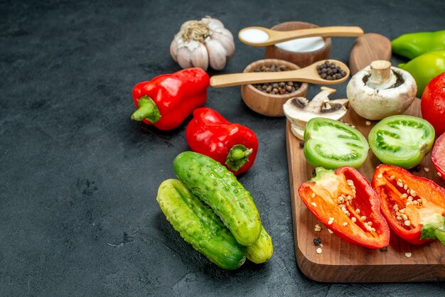 Bottom view vegetables mushrooms red and green tomatoes red bell peppers on cutting board garlic black pepper and salt in bowls wooden spoons cucumbers on dark table copy space