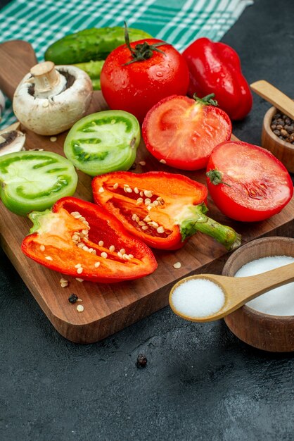 Bottom view vegetables mushrooms tomatoes bell peppers on chopping board garlic salt in bowls wooden spoons cucumbers on black table