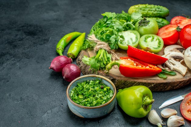 Bottom view vegetables tomatoes bell peppers cucumber greens mushroom on wood board knife bowl with greens onions on black ground