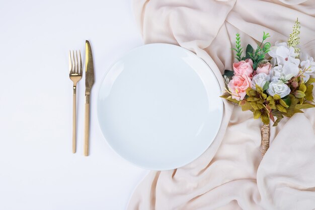 Букет цветов, тарелка и столовые приборы на белой поверхности.