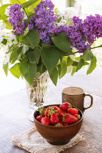 Bouquet of lilac branches in crystal vase, clay bowl with red strawberry and dark glass cup on wooden table.