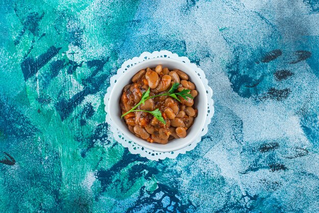 A bowl of baked beans on a coaster, on the blue table. 