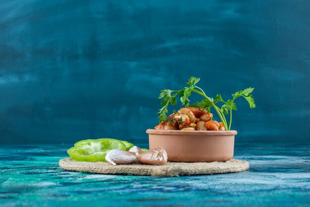 Bowl of baked beans with parsley next to garlic an pepper on a trivet , on the blue background. 