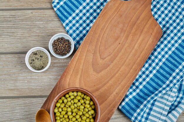 A bowl of boiled green peas with a spoon, spices, and a blue tablecloth on a wooden table.