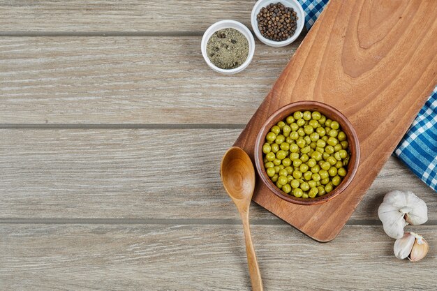 A bowl of boiled green peas with a spoon, spices, garlic, and a blue tablecloth on a wooden table.