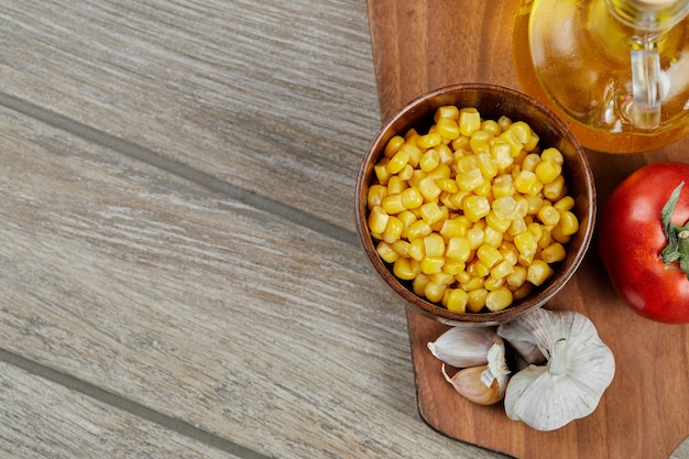 A bowl of boiled sweet corn, oil, and vegetables on a wooden board.