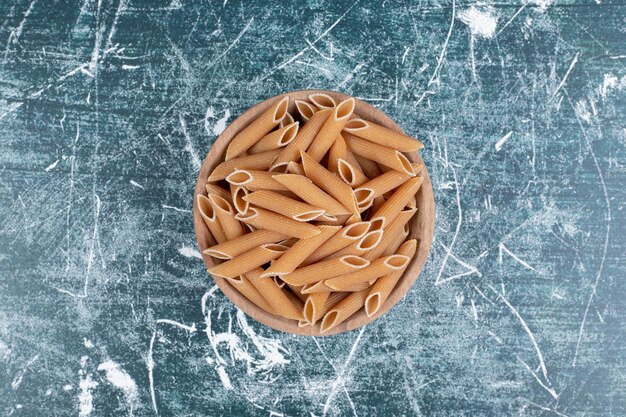 Bowl of brown raw penne pasta on blue space. 