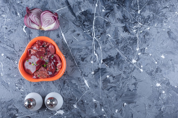 Free photo a bowl of chicken livers next to onion, salt and pepper, on the blue table.