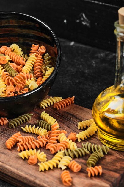 Bowl of colorful fusilli pasta and bottle of oil on dark table.