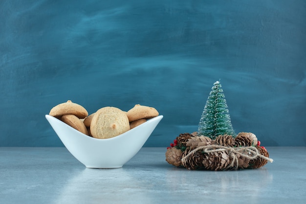 A bowl of cookie next to a tree figurine and a wreath on marble surface