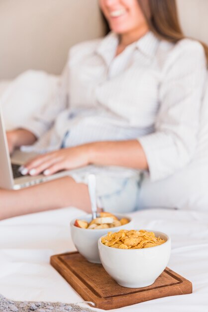 Bowl of cornflakes and fruit salad breakfast on bed with woman using laptop
