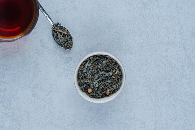 A bowl of dried tea leaves and a cup of tea with a spoon on marble.